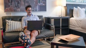 A man sitting on a couch with a laptop, a cityscape painting, a lamp, and a bed in a modern hotel room. - CCK Modern Studio Walkable to Midtown 1 Block From Marta (Atlanta)