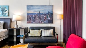 Une chambre d’hôtel moderne avec un canapé en cuir noir, un fauteuil rouge, un tableau représentant un paysage urbain accroché au mur et une table d’appoint avec une lampe.