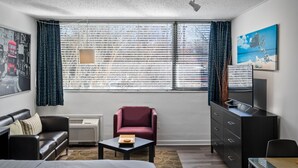Hotellihuone, jossa on sänky, punainen nojatuoli, pieni pöytä, lipasto ja suuri ikkuna, jossa on sälekaihtimet.