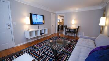 Una sala de estar moderna con un sofá, una mesa de centro y un televisor montado en la pared. Hay una zona de comedor con una mesa y sillas, y una cocina al fondo.