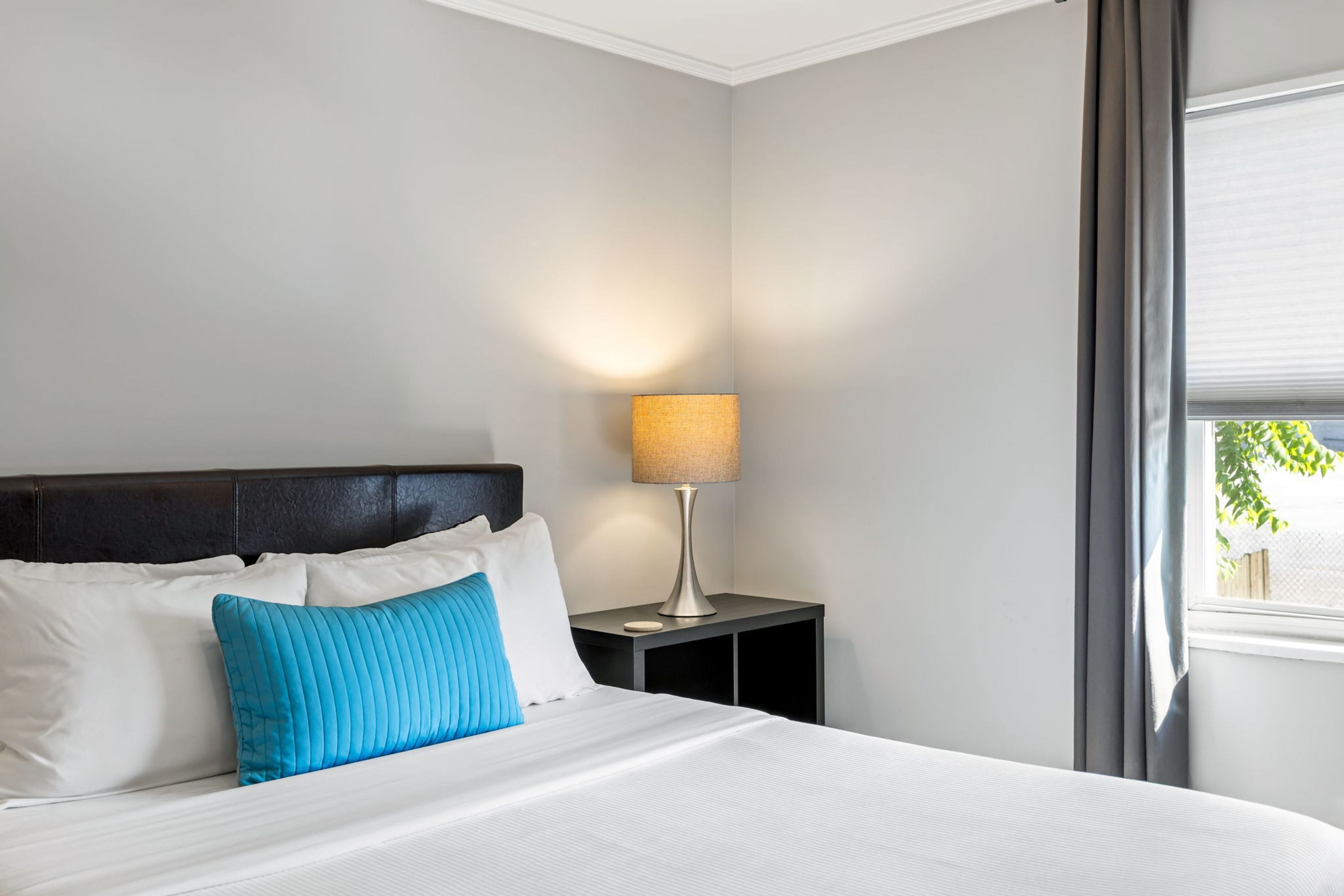 A neatly made bed with white and blue pillows, a bedside lamp, and a window with curtains.