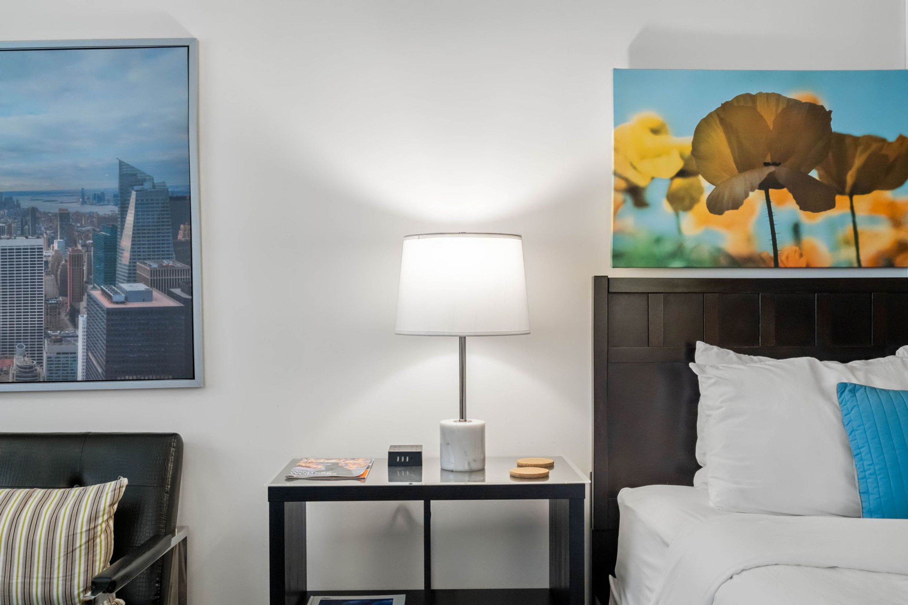Ein Hotelzimmer mit einem Bett, einem Nachttisch mit Lampe, einem Stadtbild als Gemälde und einer Blumenmotive als Gemälde.