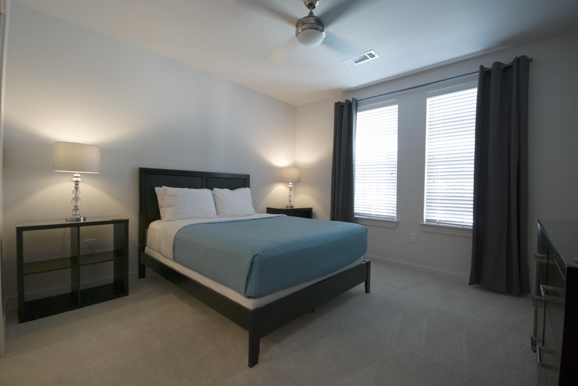 A bedroom with a bed, two bedside tables, a ceiling fan, and windows with curtains.