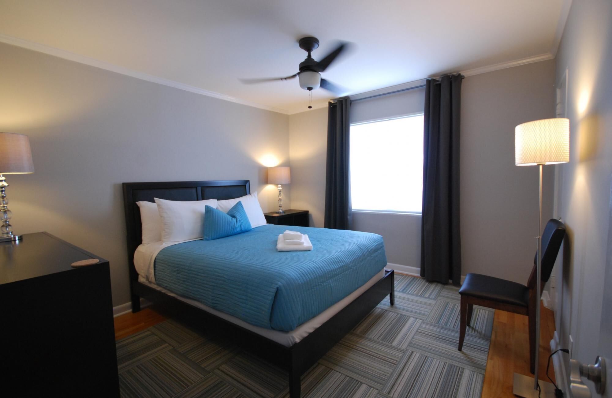 A hotel room with a bed, a ceiling fan, a nightstand with a lamp, a chair, and a window with curtains.
