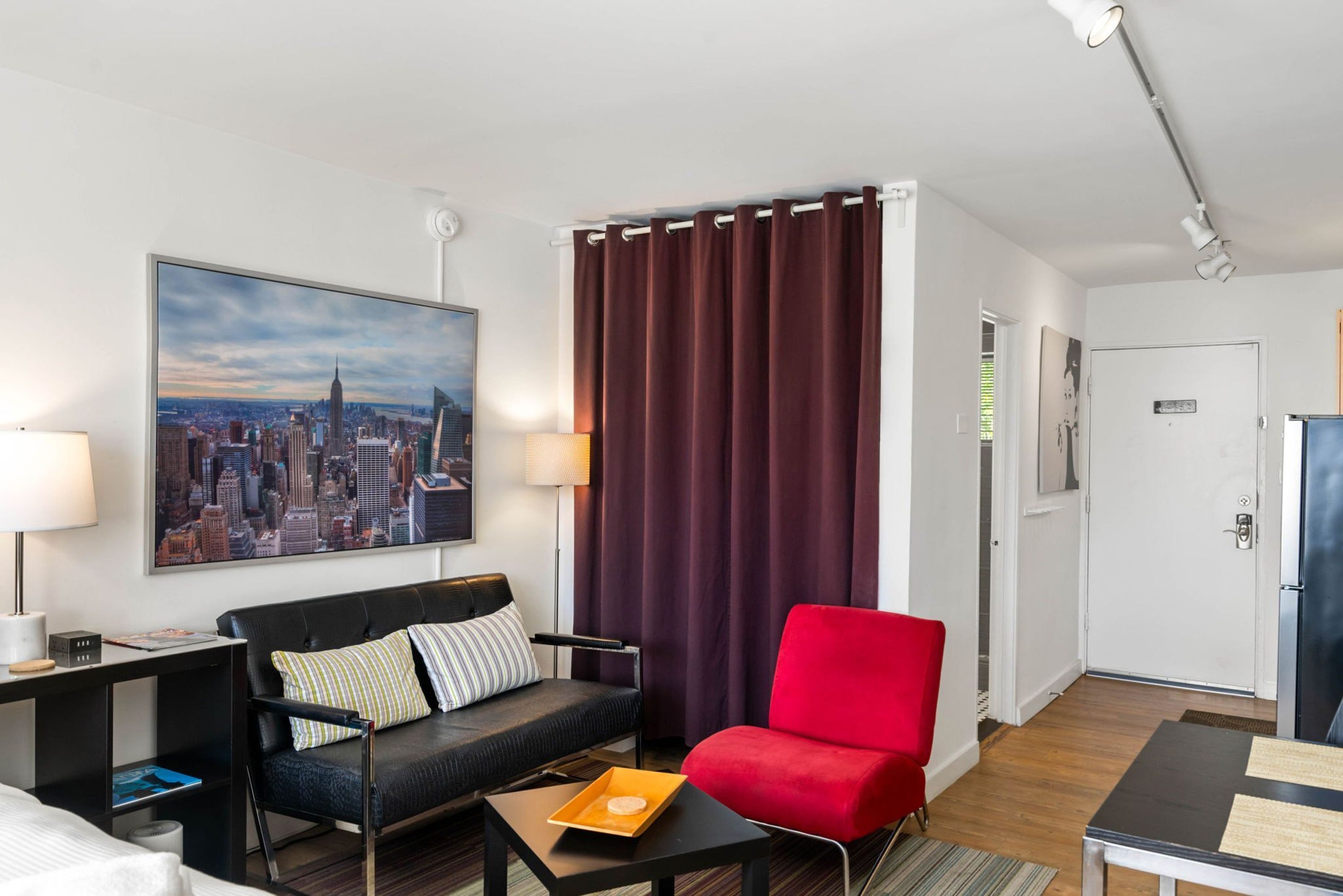 Une chambre d’hôtel moderne avec vue sur la ville, un canapé noir, un fauteuil rouge, une petite table avec un plateau, et une porte dotée d’un système de verrouillage à carte magnétique.