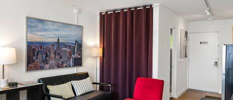 Une chambre d’hôtel moderne avec vue sur la ville, un canapé noir, un fauteuil rouge, une petite table avec un plateau, et une porte dotée d’un système de verrouillage à carte magnétique.