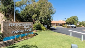 A sign for Geographe Bay Holiday Park with a brick facade, a grassy area, and a building with a red roof in the background.