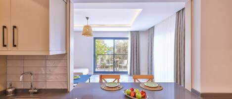 A modern kitchen with a central island, a dining area with two chairs, a large window with curtains, and a view of the outdoors.