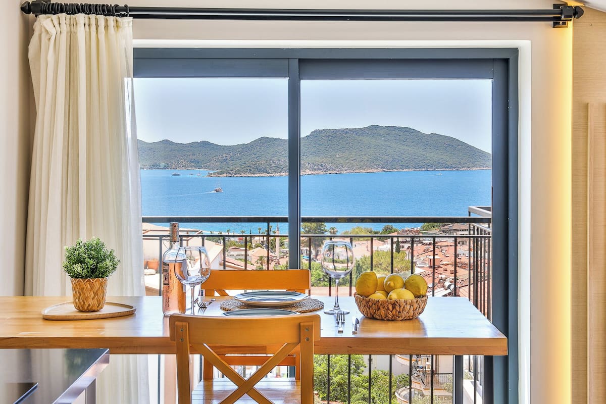 A dining area with a table set for two, a potted plant, and a view of the sea and mountains.