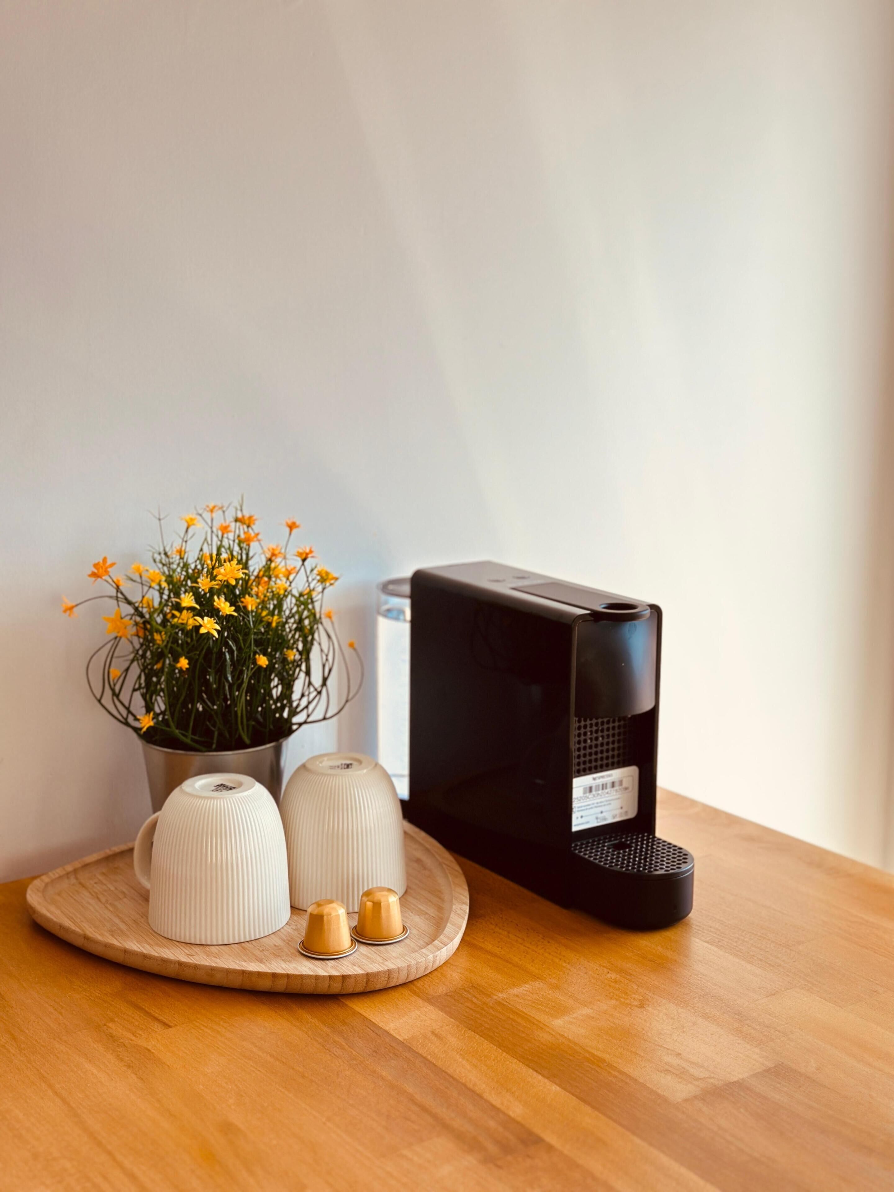 Una mesa de madera con una cafetera, una planta en maceta con flores amarillas y dos tazas blancas de cerámica.