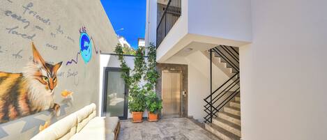 A narrow urban courtyard with a mural of a cat, a sofa, potted plants, and a staircase.