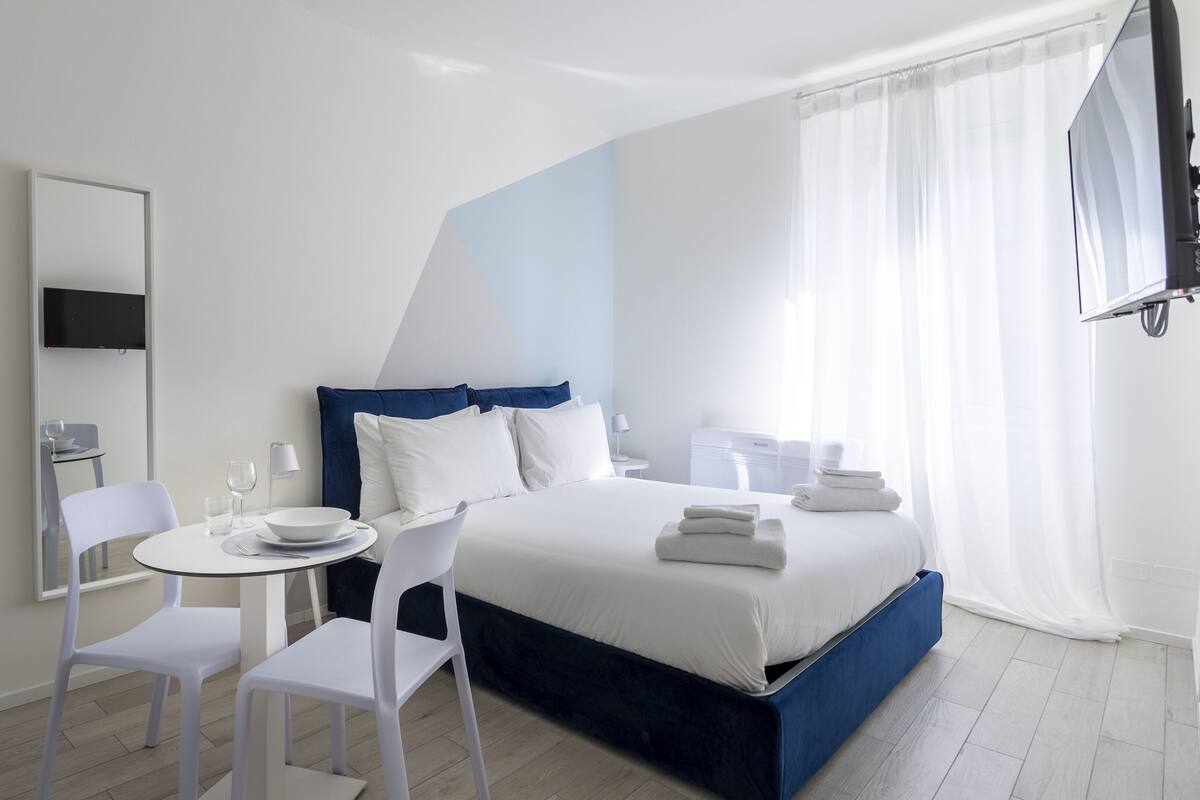 Ein modernes Hotelzimmer mit einem großen Bett, einem kleinen runden Tisch, zwei weißen Stühlen, einem Flachbildfernseher und einem großen Fenster mit durchsichtigen Vorhängen.