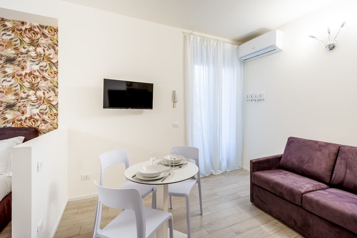 Ein modernes Wohnzimmer mit Flachbildfernseher, einem violetten Sofa, einem Esstisch mit Stühlen und einem großen Fenster mit durchsichtigen Vorhängen.