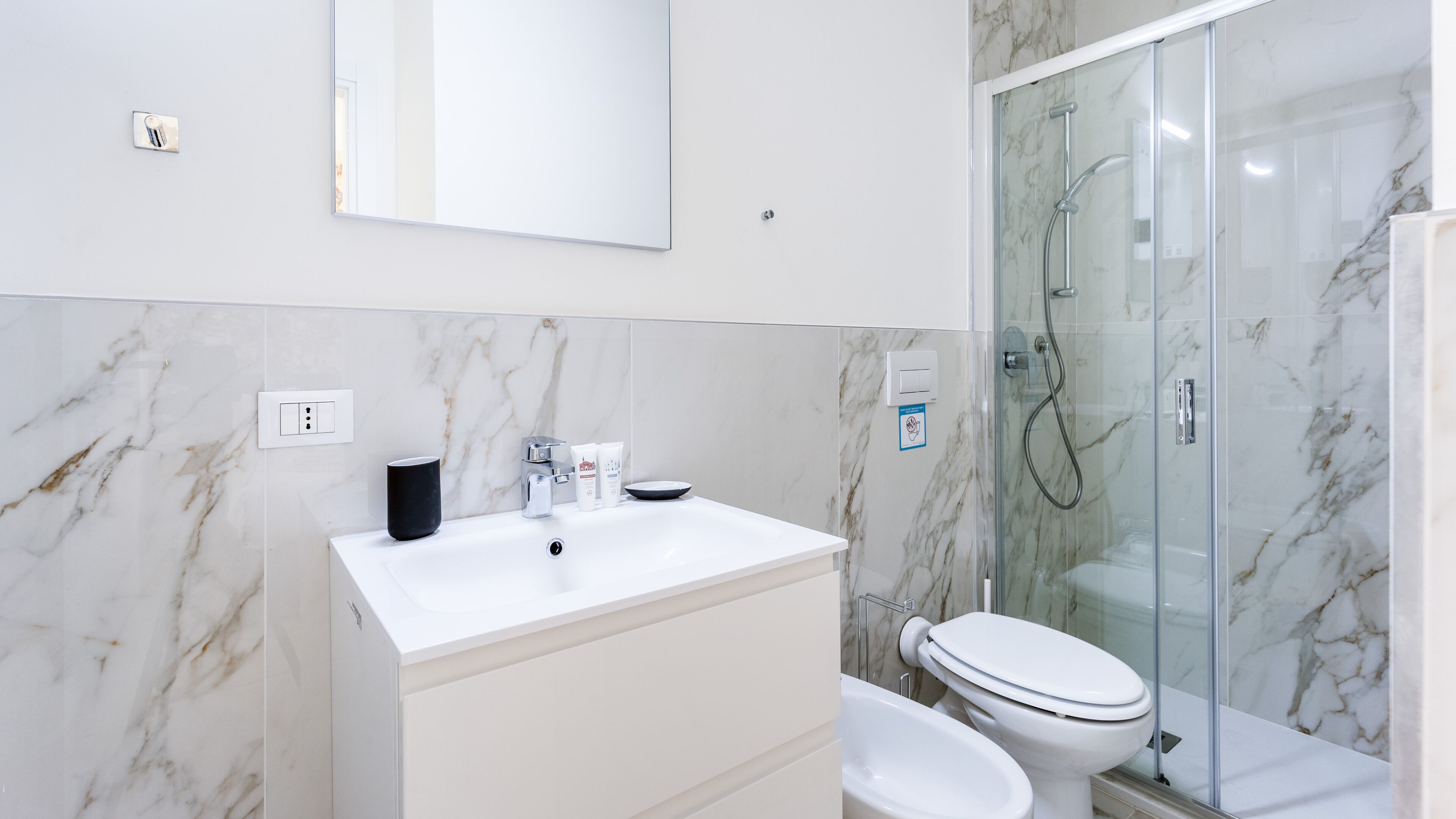 A modern bathroom with a marble backsplash, a white sink, a toilet, and a glass-enclosed shower.