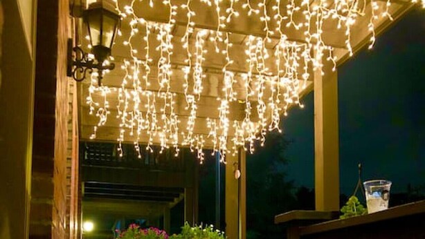 Una terraza de madera con luces colgantes, una mesa con sillas y una jardinera con flores.