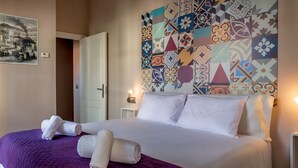A neatly made bed with white linens and a purple blanket, a patterned wall behind it, and a painting on the wall to the left. - DescubreHome Macarena Town Center (Sevilla)