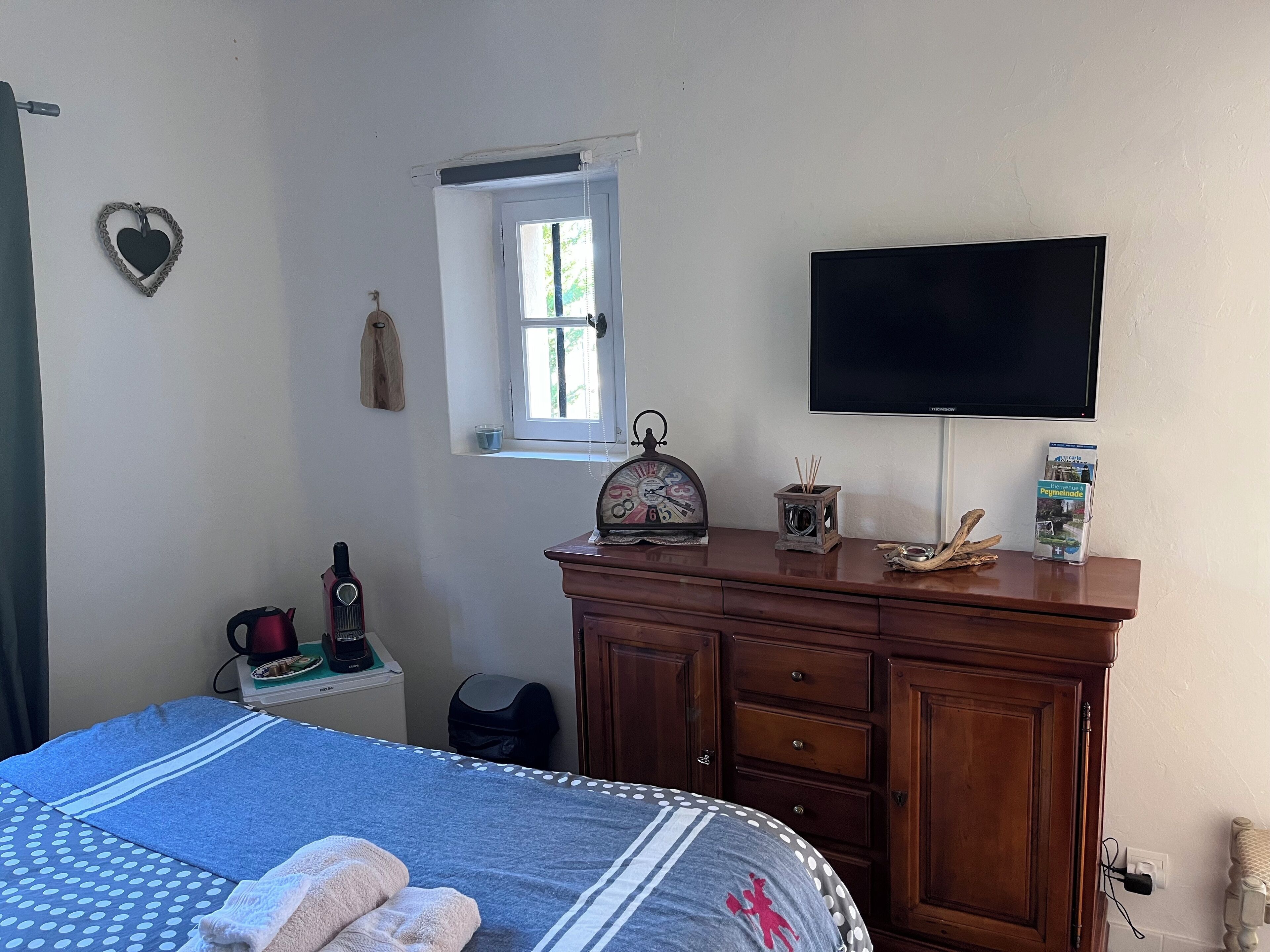 Un dormitorio con cama, cómoda, televisor, ventana y una mesita con tetera y reloj.