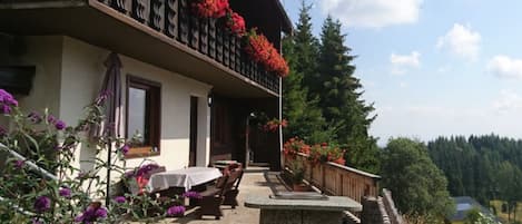 Un edificio de dos pisos con un balcón decorado con flores rojas, asientos exteriores y un muro de piedra.