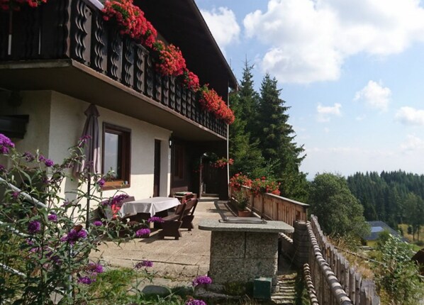 A two-story building with a balcony adorned with red flowers, outdoor seating, and a stone wall.