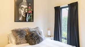 Une chambre à coucher avec un lit, une statue de Bouddha encadrée, une fenêtre avec des rideaux et une table de chevet avec une lampe.