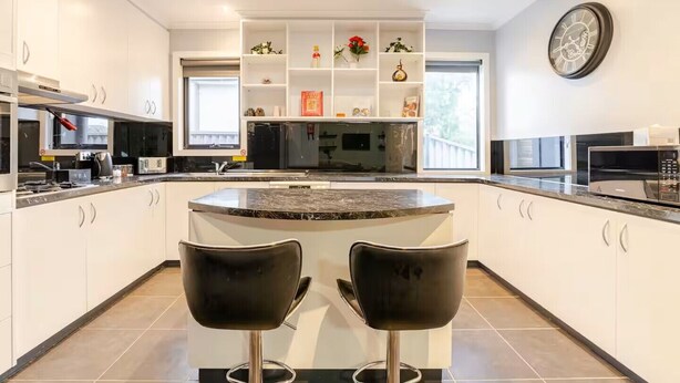 Une cuisine moderne avec des meubles blancs, un îlot central doté de deux tabourets de bar noirs, un four encastré et une horloge au mur.