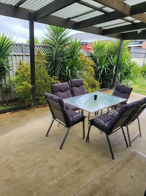 Un patio couvert doté d’une table à plateau de verre et de quatre chaises, entouré d’une végétation luxuriante et muni d’un sol en béton.
