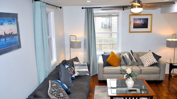 A living room with a grey sofa, a glass coffee table, and a grey couch with various cushions. There is a window with curtains, a ceiling fan, and two wall-mounted lamps. - Stylish 4-bedroom whole house with plenty of parking (Catonsville)