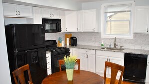 A kitchen with white cabinets, a black refrigerator, a stove, a microwave, and a dishwasher. There is a wooden dining table with chairs and a vase of flowers. - Stylish 4-bedroom whole house with plenty of parking (Catonsville)