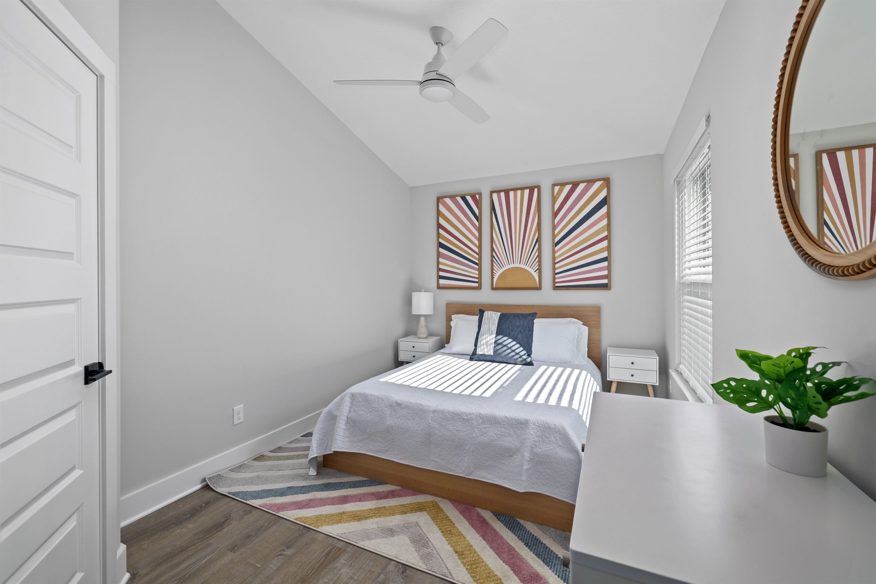 A bedroom with a bed, bedside tables, a ceiling fan, artwork on the wall, a mirror, and a potted plant.