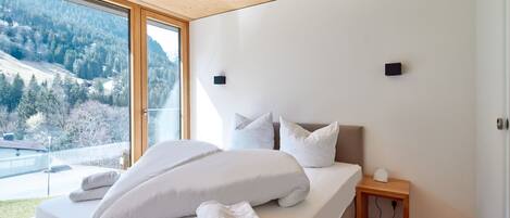A modern bedroom with a large bed, white bedding, and a view of a forested landscape through a large window.
