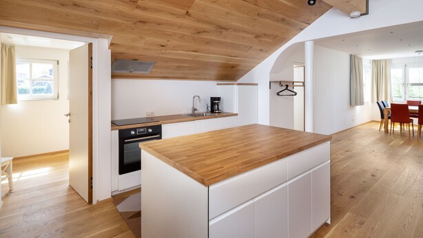 Une cuisine moderne avec un plan de travail en bois et des meubles blancs, un coin repas avec une table et des chaises rouges, et une vue à travers une porte ouverte.