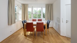Une salle à manger avec une table et des chaises en bois, de grandes fenêtres avec des rideaux et une porte blanche.