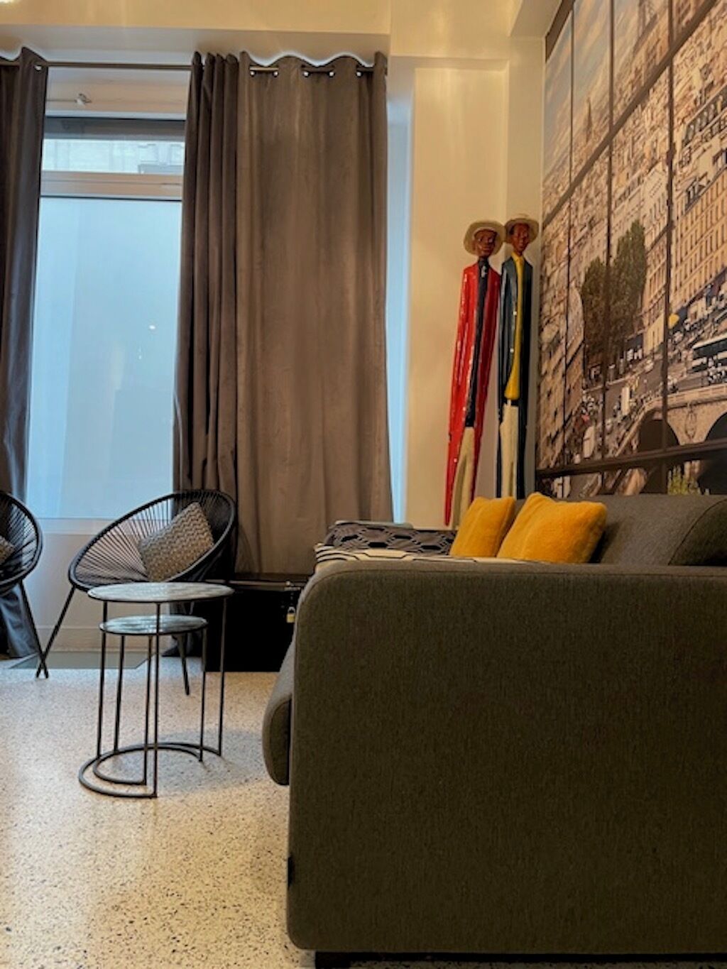 Un salon moderne avec un canapé gris, un fauteuil noir et une petite table ronde. Une grande fenêtre avec des rideaux est visible, et un grand tableau représentant un paysage urbain orne le mur.