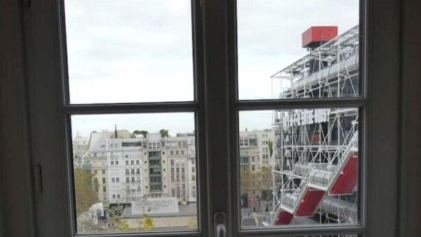 Ein Zimmer mit großem Fenster, das auf eine Baustelle mit Gerüsten und einem roten Gebäude blickt.