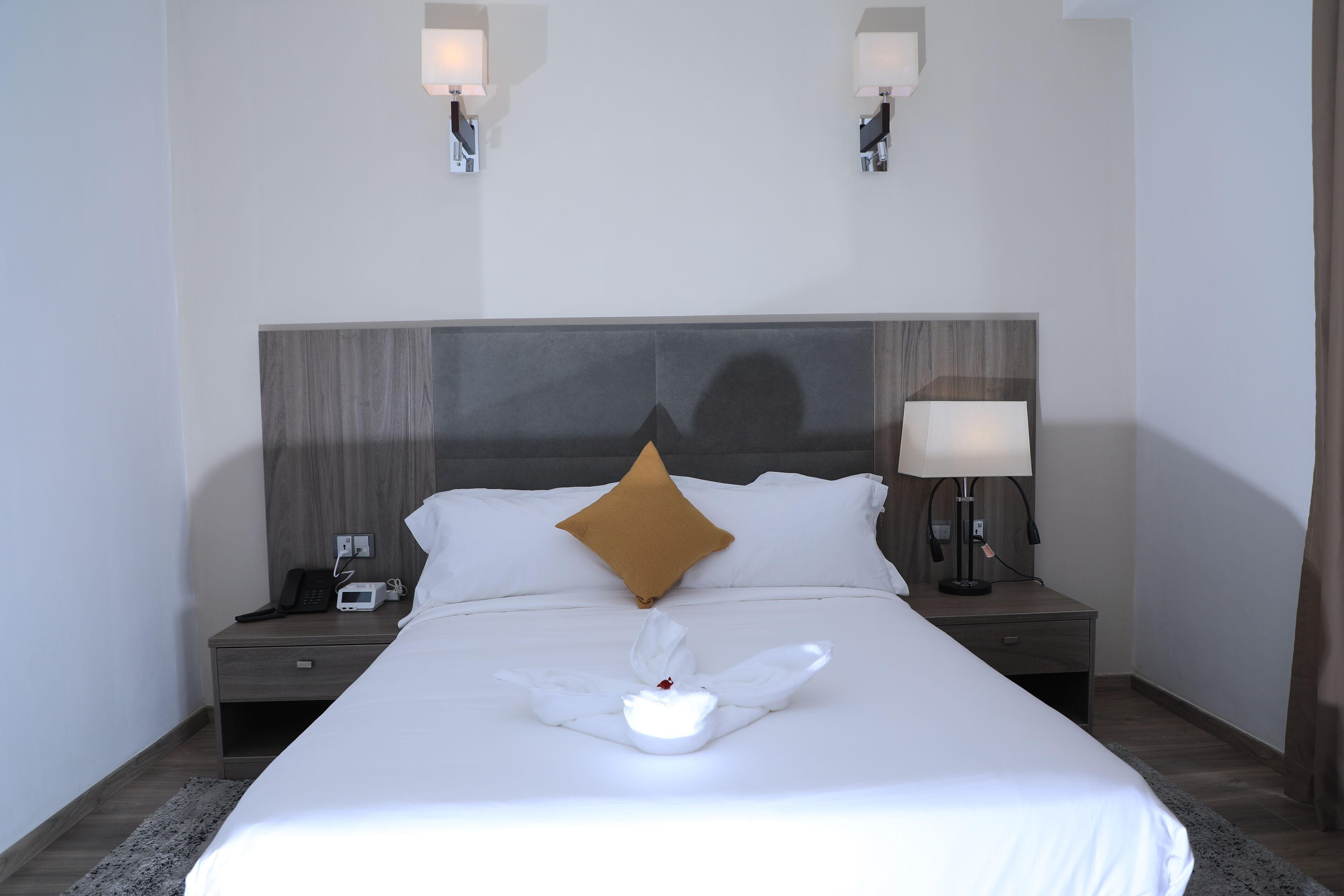 A neatly made bed with a white comforter, a brown pillow, and a white towel arrangement. There are two bedside lamps and a nightstand with a phone and a small electronic device.