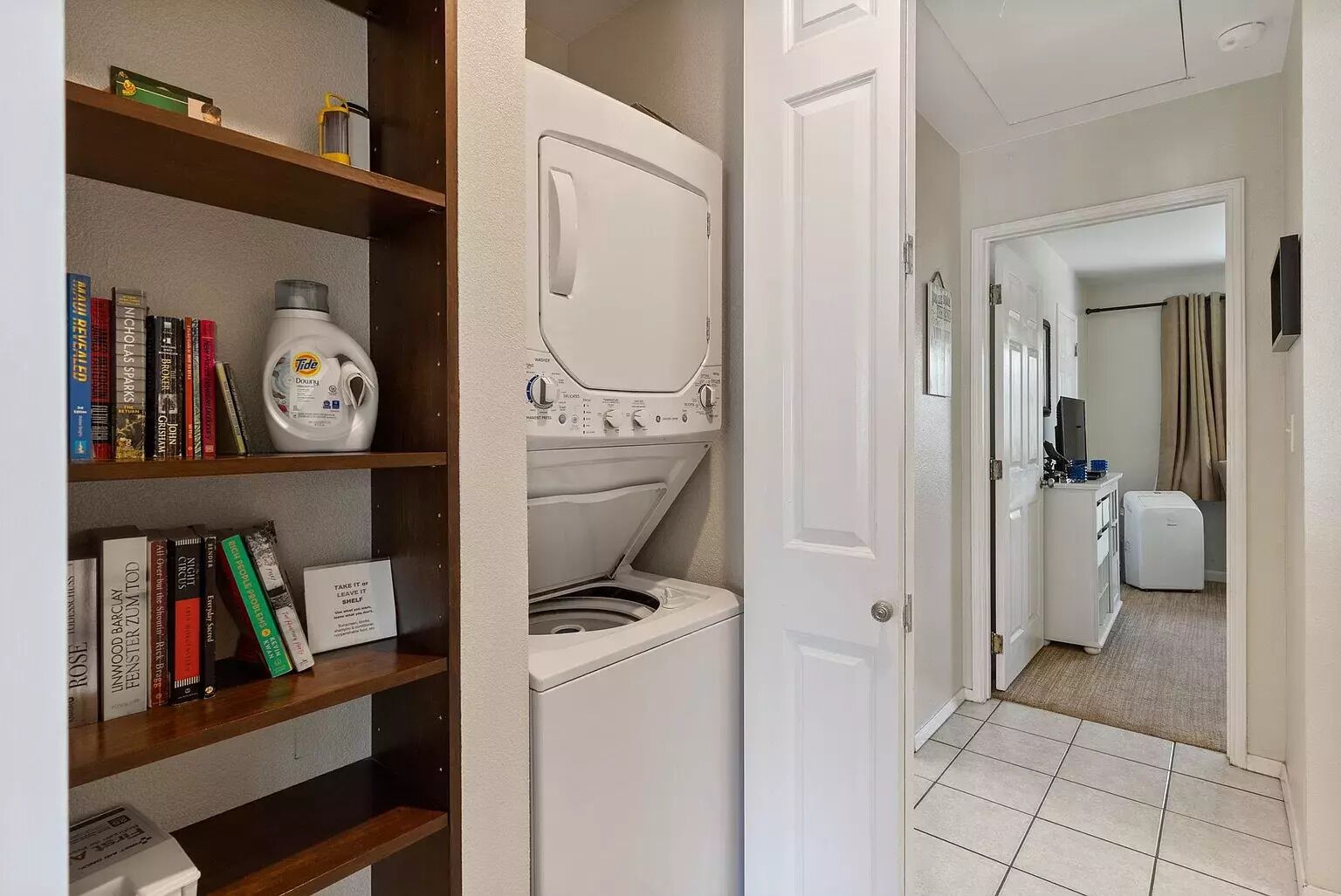A laundry room with a washing machine and dryer, a bookshelf with books, and a hallway leading to a bathroom.