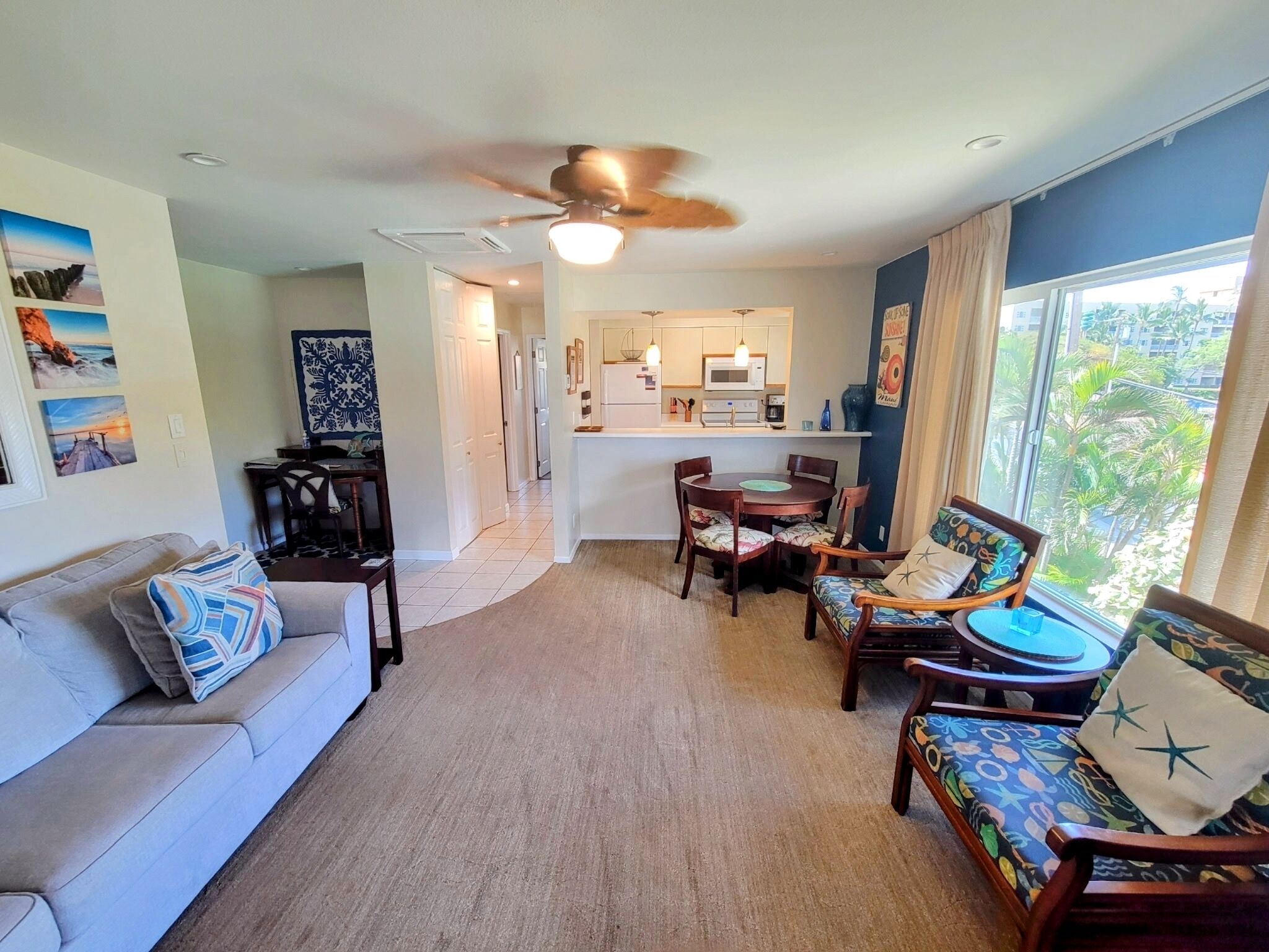 A living room with a sofa, a dining table with chairs, a kitchen area, and a ceiling fan.