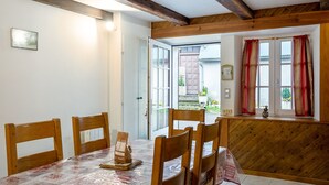 A dining area with a table set for four, wooden chairs, a wooden ceiling with exposed beams, a window with red curtains, and a door leading outside. - Gite Les Marmottes - Two Bedroom House, Sleeps 5 (Trémouille)