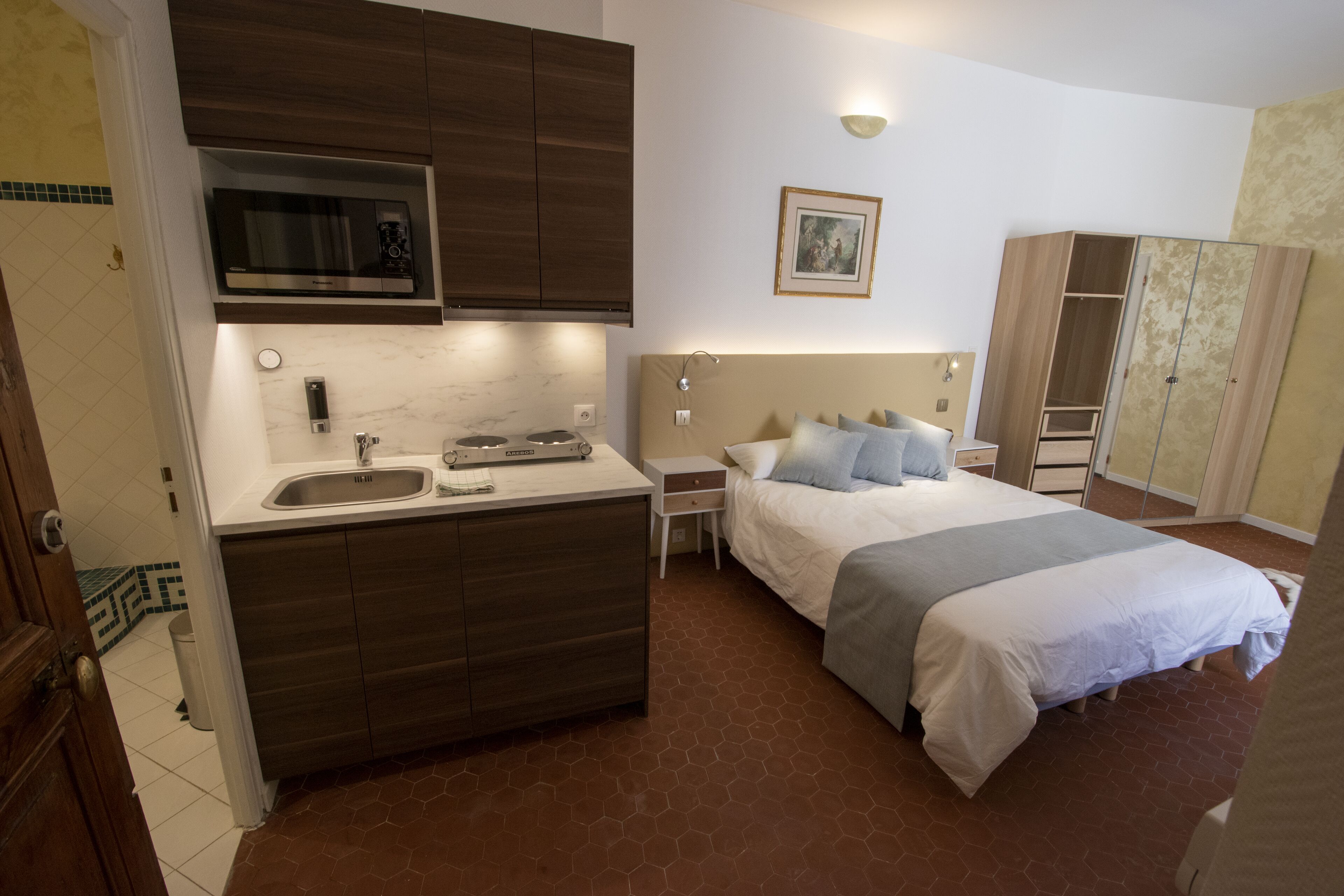 Ett kompakt hotellrum med ett litet pentry, en säng med ett grått och vitt täcke, ett sängbord och en garderob.