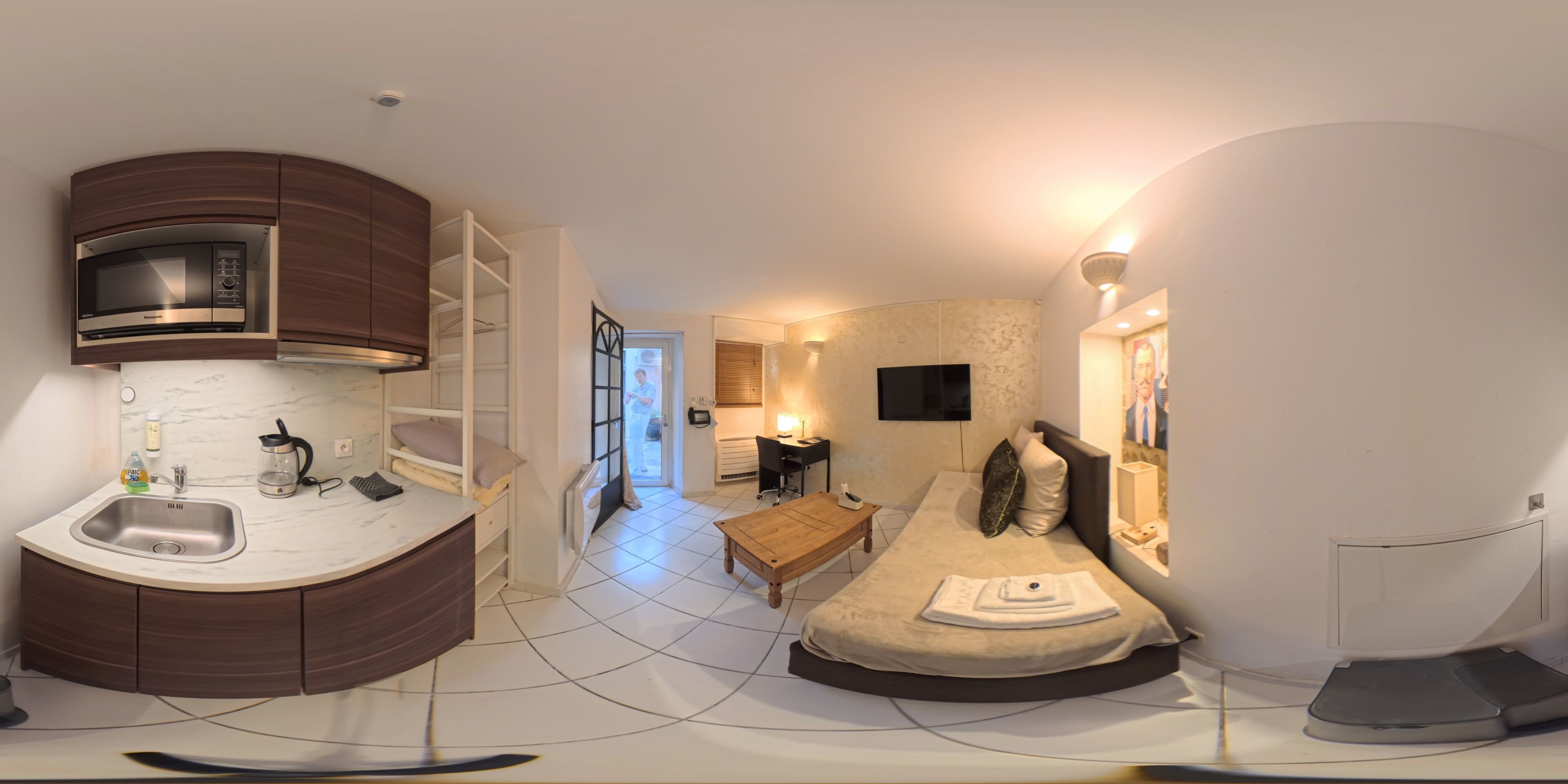 Een kleine kamer met kitchenette, een bed, een tafeltje en een televisie.