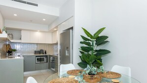 A modern kitchen with white cabinets, a stainless steel refrigerator, and a dining area with a glass table and woven placemats. - Bliss By The Ocean beautiful views, walk to beach! (Surfers Paradise)