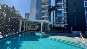A clear blue pool with lounge chairs, a covered area, and a tall building with balconies. - Bliss By The Ocean beautiful views, walk to beach! (Surfers Paradise)
