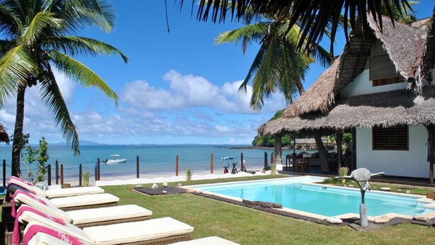 A poolside area with lounge chairs, a thatched-roof building, and a view of the ocean. - Villa Valiha Lodge (Nosy Be)