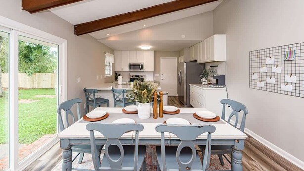 A dining area with a table set for six, surrounded by chairs, in a modern kitchen with white cabinets and a large window. - Charming 3-Bedroom Retreat with Private Yard (Charleston)