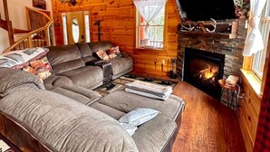 A cozy log cabin interior with a large sectional sofa, a fireplace, and a television mounted on the wall.