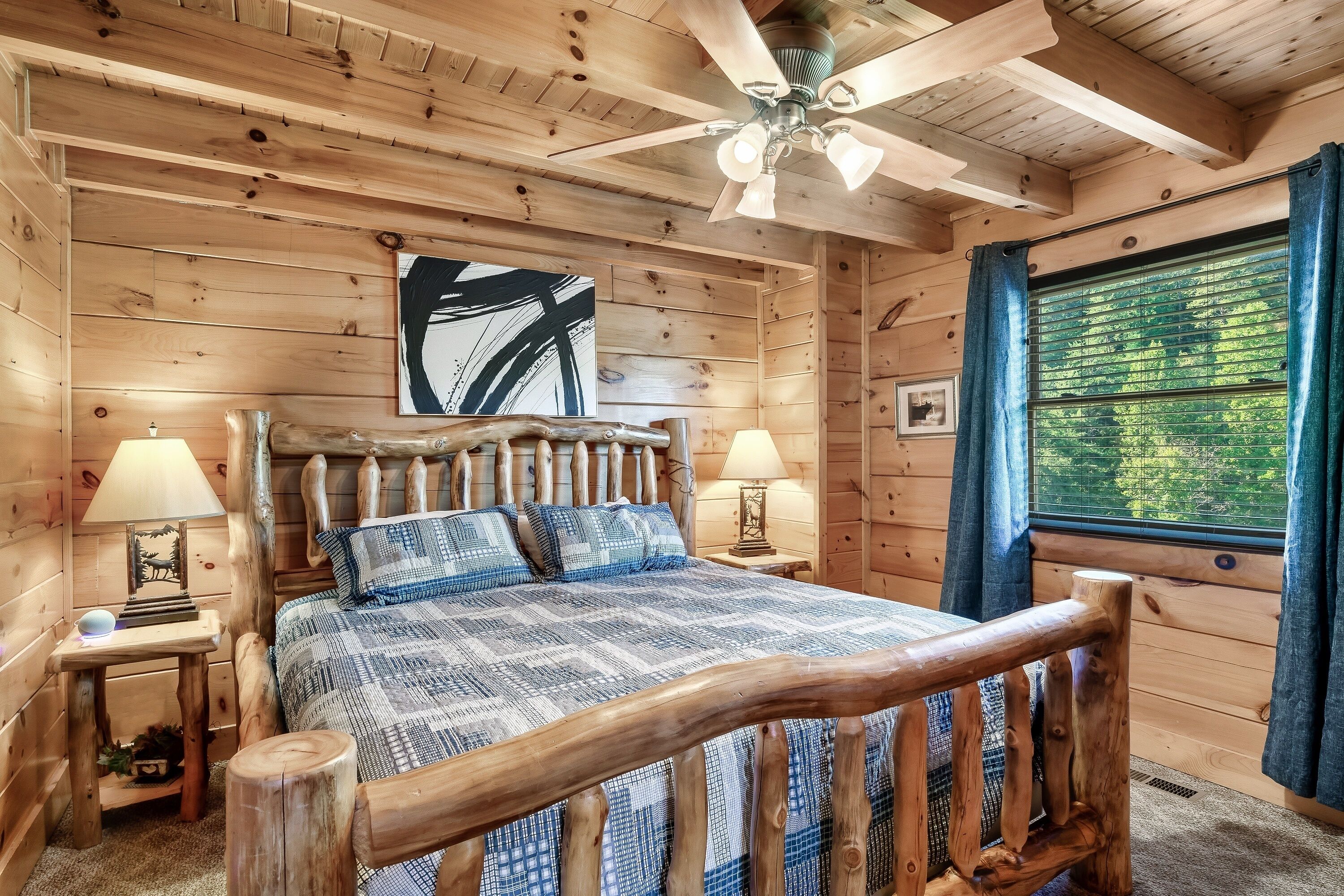 Une chambre dans une cabane en bois, avec un lit, deux lampes de chevet, un ventilateur de plafond et une fenêtre avec des rideaux.