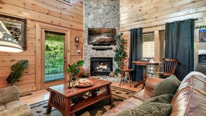 Un salon chaleureux avec une cheminée en pierre, des murs en bois, des meubles en cuir et une grande fenêtre donnant sur la forêt.