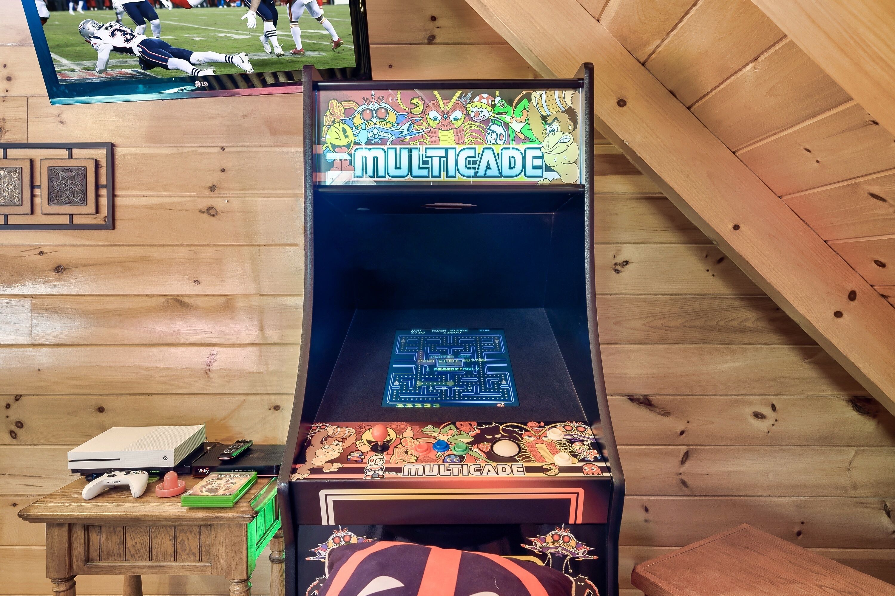 Una habitación con paneles de madera, una máquina arcade Multicade, una consola de videojuegos blanca y un televisor montado en la pared.