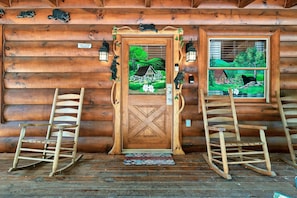 A wooden cabin with a wooden door, two rocking chairs, and a window with a painting of a house and trees.