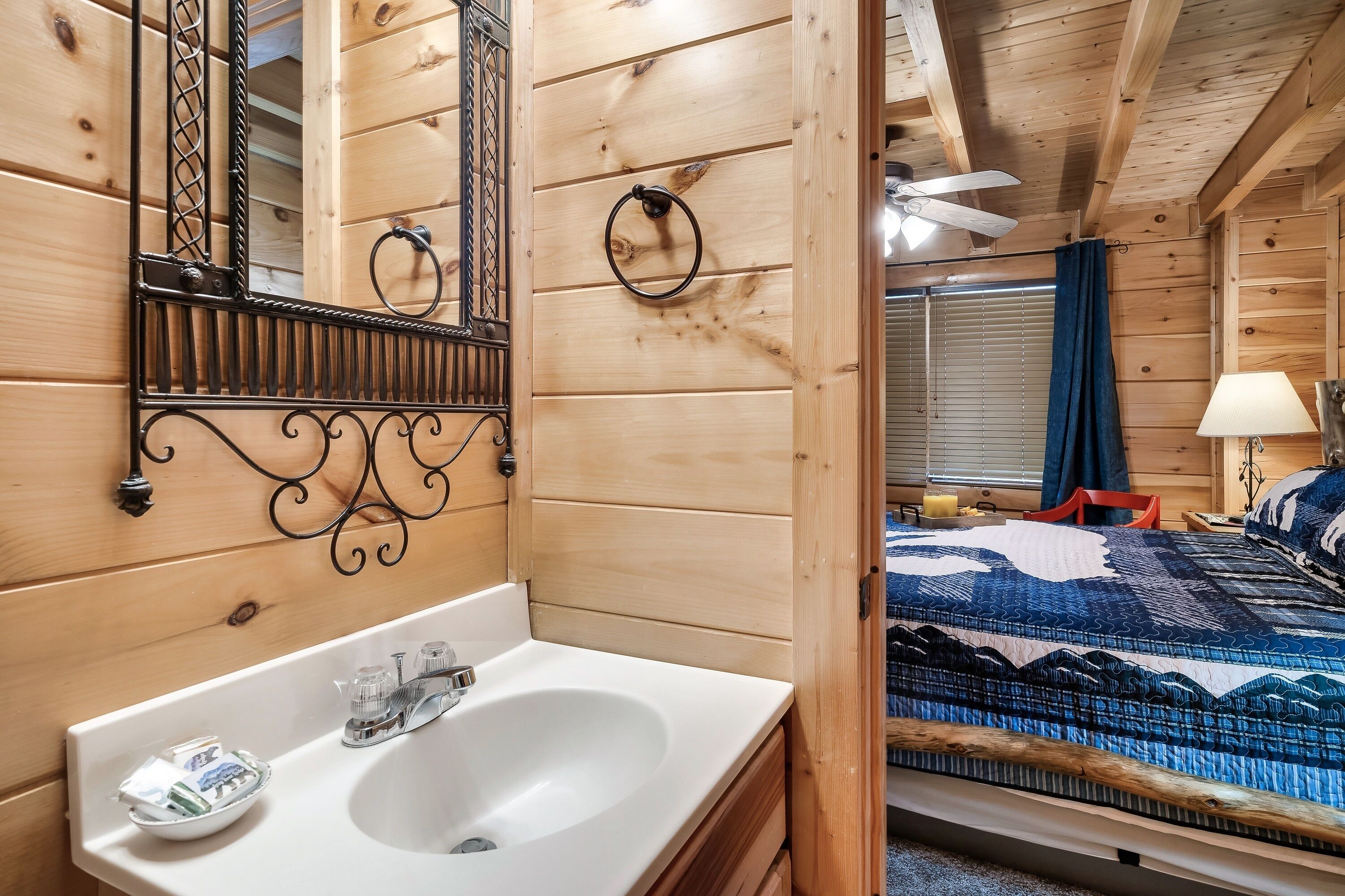 Ein Holzblockhaus-Innenraum mit einem weißen Waschbecken, einem Spiegel mit Zierrahmen, einem Bett mit blau gemustertem Bettbezug und einem Deckenventilator.